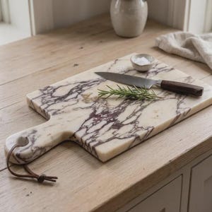 May include: A rectangular marble serving board featuring white and deep purple veining, a handle with a brown leather cord, a stainless steel knife with a dark wooden handle, a small ceramic bowl of salt, and a sprig of fresh green rosemary.