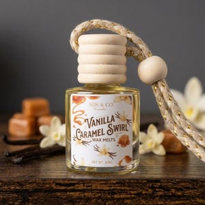 May include: A small glass bottle filled with a clear liquid, topped with a ribbed wooden cap and a braided beige cord. The label features the text NIN & CO. CANDLES VANILLA CARAMEL SWIRL WAX MELTS NET WT. 8 MIL, with illustrations of vanilla flowers and caramel.