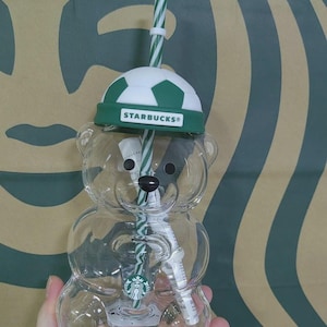 May include: A clear, bear-shaped plastic tumbler featuring a green and white soccer ball-patterned lid with the Starbucks logo text. The cup includes a green and white striped straw and a small circular Starbucks siren logo on the bear's chest.
