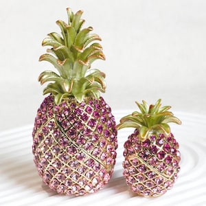 May include: Two pineapple-shaped trinket boxes featuring a gold-tone metal frame encrusted with sparkling pink crystals. Each pineapple has a textured green metal top representing the leaves, with a hinged design for opening the main body.