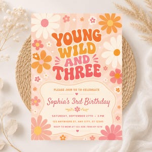 Op de afbeelding: Een rechthoekige verjaardagsuitnodigingskaart met de tekst YOUNG WILD AND THREE in een oranje en roze retro-lettertype. Het ontwerp bevat oranje, roze en witte madeliefjes op een golvende perzikkleurige achtergrond met details voor de 3e verjaardag van Sophia.