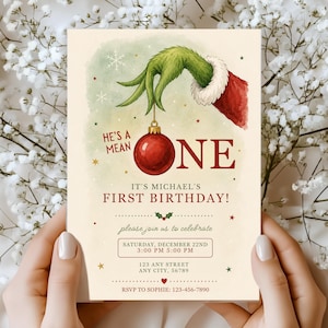 May include: A rectangular birthday invitation card featuring a green furry hand in a red sleeve holding a red Christmas ornament. The text reads: HE'S A MEAN ONE IT'S MICHAEL'S FIRST BIRTHDAY! SATURDAY, DECEMBER 22ND 3:00 PM 5:00 PM 123 ANY STREET ANY CITY, 56789.