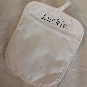 May include: A white quilted pot holder with a pocket featuring the name Luckie embroidered in black script lettering. The square-shaped kitchen accessory includes a small fabric loop at the top corner for hanging and has a smooth white border.