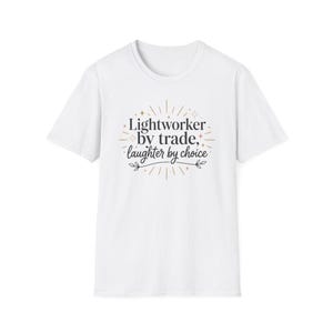 May include: A white short-sleeve t-shirt featuring the black text Lightworker by trade, laughter by choice. The design is surrounded by thin gold lines, small star icons, and a decorative flourish with leaves at the bottom center of the graphic.