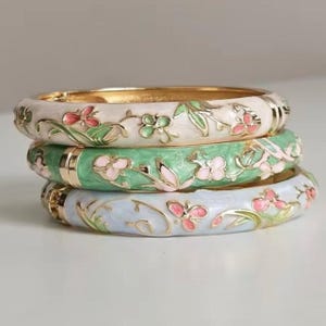 May include: A stack of three metal bangle bracelets featuring enamel floral and butterfly motifs. The top bangle is cream, the middle is light green, and the bottom is light blue. Each bracelet has gold-toned metal accents and hinges.