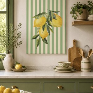 Op de afbeelding: Een rechthoekige wandkunstprint met een aquarelillustratie van drie gele citroenen met groene bladeren, gecentreerd op een achtergrond van verticale saliegroene en crèmekleurige strepen. Het kunstwerk hangt aan een muur boven een aanrecht.