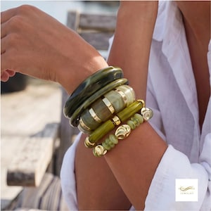 Pode incluir: Um conjunto de cinco pulseiras em vários tons de verde-azeitona e sálvia. O conjunto inclui dois braceletes lisos verde-escuros, um bracelete segmentado verde e dourado, uma pulseira de tubo verde-azeitona e uma pulseira de contas com detalhes dourados.
