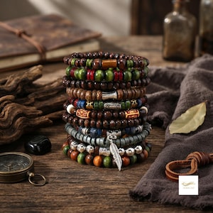 Op de afbeelding: Een stapel van tien kralenarmbanden met diverse materialen, waaronder donkerbruine, groene, rode en blauwe houten kralen, metalen tussenstukjes en een zilverkleurige bedel in de vorm van een veer, verticaal gerangschikt in één kolom.