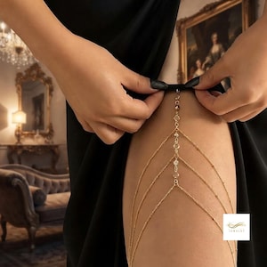 Può includere: Una catena da coscia in metallo color oro caratterizzata da molteplici catenine sottili drappeggiate e piccoli cristalli circolari trasparenti. Il gioiello è fissato in alto con un anello e si estende verso il basso in un design a strati.