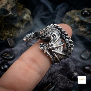 Puede incluir: Un anillo de tono plateado con forma de dragón enroscado con escamas detalladas, cuello largo y alas plegadas. El metal tiene un acabado oscuro y oxidado en las hendiduras para resaltar la textura del cuerpo y las alas del dragón.