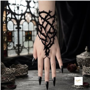 Könnte beinhalten: Ein schwarzer Handschmuck mit einem Gitterdesign aus ineinander verschlungenen dornigen Ranken. Das Stück bedeckt den Handrücken und ist mit einer dünnen Kette mit einem Ring am Mittelfinger verbunden, was eine strukturierte, käfigartige Optik erzeugt.