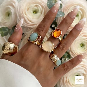 Peut inclure: Une collection de cinq bagues dorées portées sur les doigts. Les bagues présentent des designs variés, dont une large bague en or martelé, une bague avec une pierre ovale bleu clair, une bague avec une pierre orange translucide, une bague avec une pierre crème et une bague texturée avec une petite pierre bleu clair.