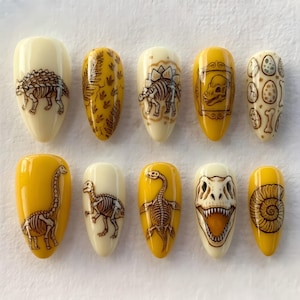 May include: A set of ten almond-shaped press-on nails featuring yellow and cream backgrounds with detailed brown line art illustrations of dinosaur skeletons, fossils, eggs, and prehistoric plants arranged in two horizontal rows.