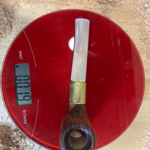 May include: A wooden smoking pipe with a dark brown grain, a gold-colored metal band, and a translucent white stem. The pipe is positioned on a red digital scale that displays a weight of 0.19 ounces, showing the pipe's mass in a clear, objective manner.