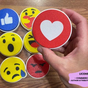 May include: A collection of circular coasters featuring various social media reaction icons and emojis, including a blue thumbs-up, a red heart, and several yellow faces expressing emotions like surprise, laughter, sadness, and anger with a textured finish.