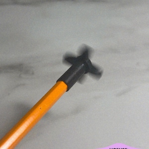 May include: A bright orange pencil featuring a black, four-bladed propeller attachment fixed to the eraser end. The propeller appears to be in motion, creating a blurred effect around the black plastic blades attached to the pencil shaft.
