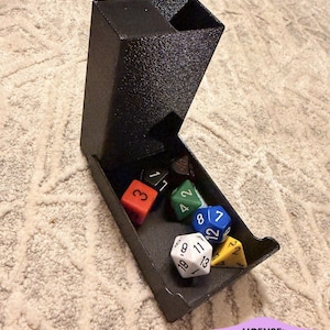 May include: A black textured dice tower with an open tray containing several multi-sided gaming dice in various colors including orange, green, blue, white, and yellow. The tower features a vertical chute and a front-facing landing area for dice.