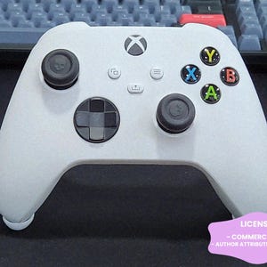 May include: A white wireless video game controller featuring two black analogue sticks with skull icons, a black directional pad, and four coloured action buttons labelled X, Y, A, and B. The controller includes a central Xbox logo button and three small menu buttons.