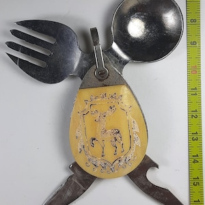Puede incluir: Un utensilio de camping multifunción con tenedor, cuchara, cuchillo y abrebotellas de plata unidos a un mango color crema con un grabado de un escudo de ciervo negro. Una cinta métrica amarilla muestra que mide unos 23 cm.