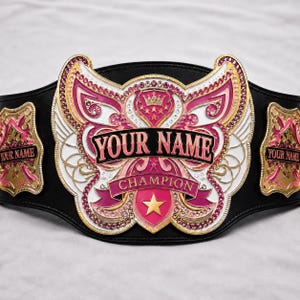 Könnte beinhalten: Ein Meisterschaftsgürtel aus schwarzem Leder mit einer großen, verzierten Mittelplatte aus rosa und weißem Emaille, goldenen Akzenten und eingebetteten Kristallen. Die Platte zeigt den Text YOUR NAME und CHAMPION sowie seitliche Platten mit YOUR NAME.