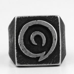 May include: A square-faced signet ring crafted from dark, oxidized metal. The face of the ring features a deeply recessed, textured background with a raised, polished silver-toned spiral symbol centered on the square surface.