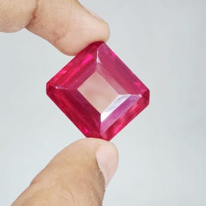 Op de afbeelding: Een vierkant geslepen, levendig rode edelsteen met een gefacetteerd oppervlak dat licht reflecteert. De steen heeft scherpe, strakke randen en een transparante, dieprode kleur, met een gepolijste afwerking die de vorm benadrukt.