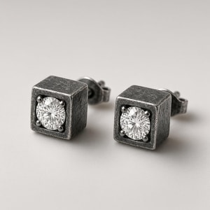 May include: A pair of stud earrings featuring a square, dark gray, textured metal setting. Each earring holds a single, round, clear, faceted gemstone centered within the cube. The earrings include standard post backs for secure attachment.