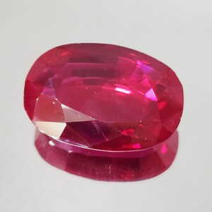 May include: A vibrant, oval-cut gemstone with a deep, saturated red color. The stone features numerous triangular and trapezoidal facets that create a brilliant, reflective surface, showcasing its translucent quality and intense crimson hue.