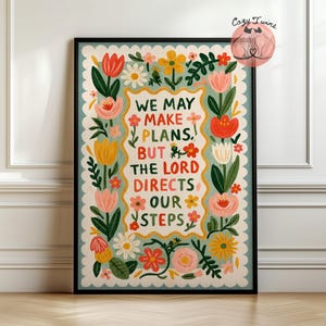 Può includere: Una stampa artistica incorniciata con sfondo azzurro, bordo smerlato e motivo floreale. Il testo centrale recita: WE MAY MAKE PLANS, BUT THE LORD DIRECTS OUR STEPS. Il design include fiori di colore rosso, giallo, rosa e bianco.