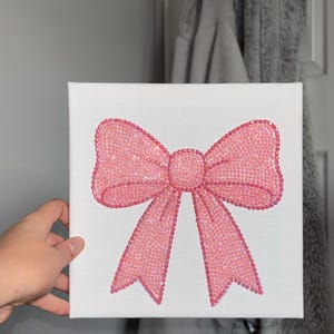 May include: A square white canvas featuring a central bow design composed of numerous small, shimmering pink rhinestones. The bow is depicted with a central knot and two long, downward-pointing tails, all covered in the reflective pink gems.