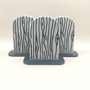 May include: Three light gray, arched, tombstone-shaped objects featuring a black, hand-drawn wood grain pattern. Each piece is mounted on a dark gray, flat, rectangular base, arranged in a cluster with one piece positioned in the front center.