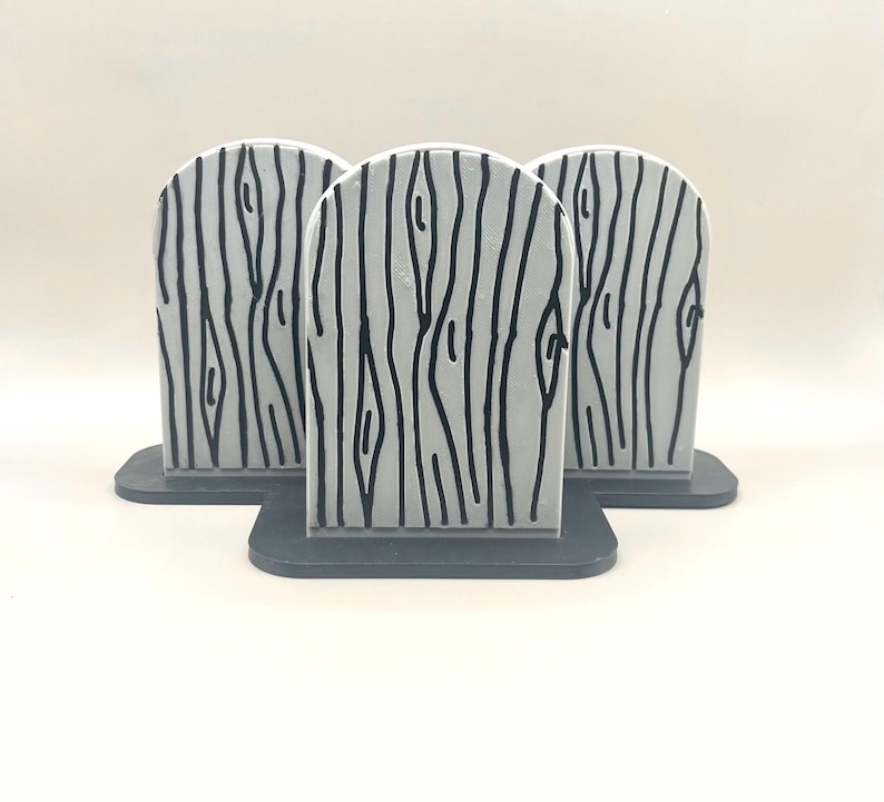 May include: Three light gray, arched, tombstone-shaped objects featuring a black, hand-drawn wood grain pattern. Each piece is mounted on a dark gray, flat, rectangular base, arranged in a cluster with one piece positioned in the front center.