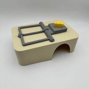 May include: A beige rectangular structure with a rounded archway opening on the side. On top sits a grey mechanical trap mechanism featuring a spring-loaded bar and a small, yellow, triangular piece of cheese with holes resting on a grey platform.
