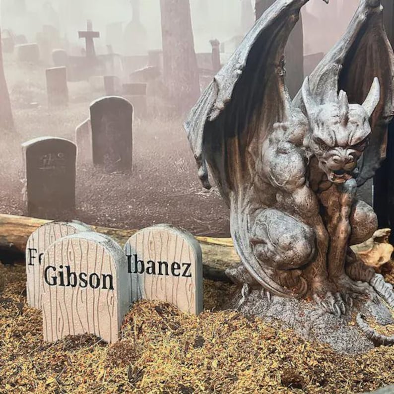 May include: A grey stone-colored gargoyle statue with large wings crouched next to three small, light-colored tombstone-shaped markers. The markers feature wood grain textures and the black printed text labels Gibson and Ibanez.