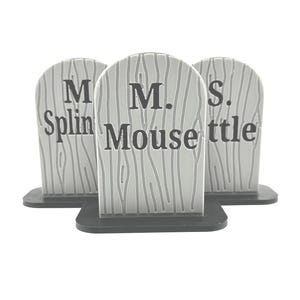 May include: Three gray, arched, tombstone-shaped decorations with a wood grain texture and black bases. Each piece features black text: the center one reads M. Mouse, while the flanking pieces display partial text reading M. Splin and S. ttle.