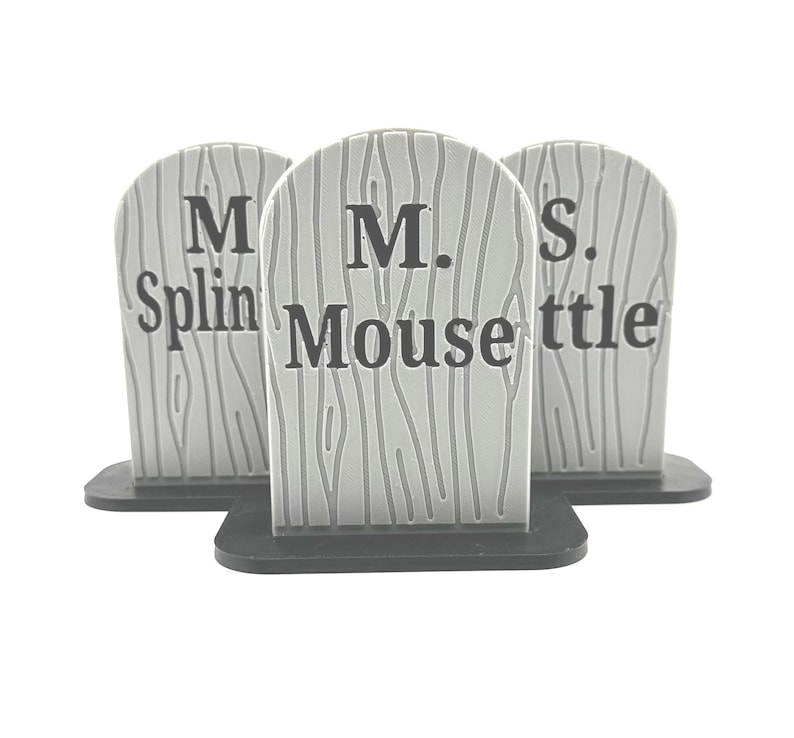 May include: Three gray, arched, tombstone-shaped decorations with a wood grain texture and black bases. Each piece features black text: the center one reads M. Mouse, while the flanking pieces display partial text reading M. Splin and S. ttle.
