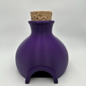 May include: A purple, bulbous, bottle-shaped container with a smooth matte finish. The top features a textured, light tan, cylindrical stopper. The base of the container has a wide, arched opening cut into the front, creating a hollow interior.