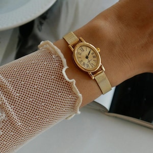 Può includere: Un orologio da polso color oro con cassa ovale, quadrante color crema, numeri romani neri come indicatori delle ore e lancette nere sottili. L'orologio include un cinturino in maglia metallica color oro fine con un meccanismo di chiusura.