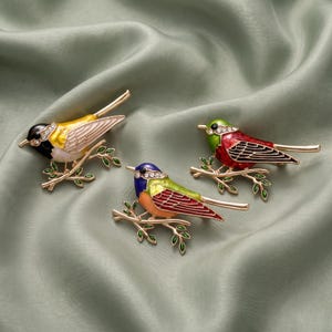 May include: Three bird-shaped brooches crafted from gold-tone metal with colorful enamel detailing. Each brooch features a bird perched on a branch with green leaves, accented by small clear rhinestones along the head and wing areas.