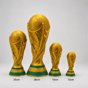 May include: Four gold-coloured FIFA World Cup trophy replicas of varying sizes, featuring a globe supported by stylised figures on a green base. The heights are labelled as 25cm, 36cm, 15cm, and 12cm. The text FIFA WORLD CUP is embossed on the green base.