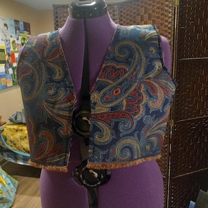 May include: A sleeveless vest garment featuring a dark blue fabric base with an intricate, multi-colored paisley pattern in shades of orange, yellow, and teal. The vest has a raw, fringed hemline along the bottom edge and is displayed on a purple mannequin.