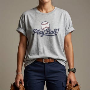 May include: A heather grey short-sleeved t-shirt featuring a graphic of a white baseball with red stitching and the dark blue text Play Ball! written in a cursive script font across the chest. The shirt has a crew neckline and rolled sleeves.