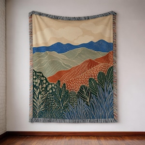 May include: A woven throw blanket featuring a landscape illustration with a cream sky, blue and sage green mountains, and terracotta hills. The foreground displays dark green foliage and blue plants, finished with a multicolored fringe border.