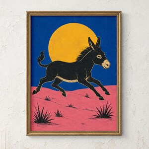 May include: A framed art print featuring a black donkey running across a pink desert landscape with small black plants. The background consists of a large yellow sun set against a deep blue sky, all enclosed within a thin metallic gold frame.
