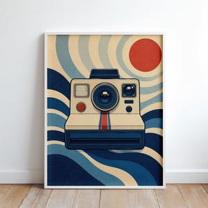 May include: A framed wall art print featuring a cream-colored instant camera with a central red and blue vertical stripe. The background displays a graphic pattern of wavy blue and cream lines radiating from a solid red circle in the upper right corner.