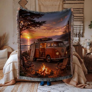 May include: A woven throw blanket featuring a sunset beach scene with a vintage orange and white camper van, a surfboard leaning against it, two folding chairs, and a campfire. String lights hang above the van under a colorful evening sky.