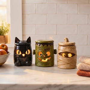Puede incluir: Tres botes de cerámica con temática de Halloween y tapas, iluminados desde el interior. A la izquierda un gato negro, en el centro un monstruo verde con costuras y a la derecha una momia beige. Cada uno tiene ojos y bocas recortados brillantes.