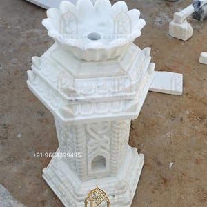 May include: A white marble Tulsi Vrindavan featuring a lotus-shaped top basin with a central hole, a tiered octagonal base, and intricate carved architectural details on the sides. The structure includes the text Marble Tulsi Vrindavan and +91-9664399495.