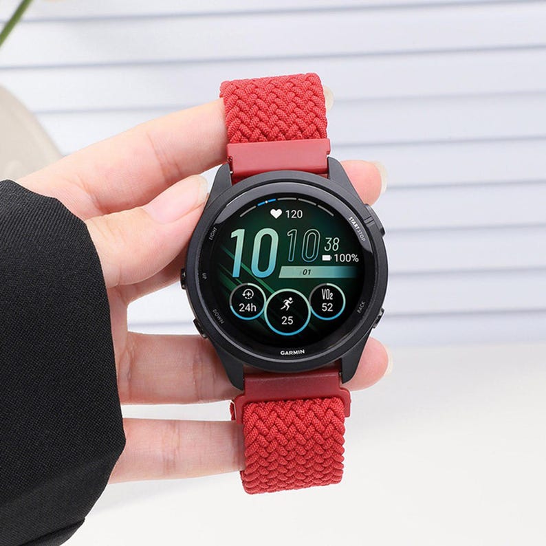 May include: A Garmin smartwatch with a black circular case and a bright red braided nylon watch band. The digital display shows the time as 10:10, a heart rate of 120, a battery level of 100%, and various activity tracking icons and data points.