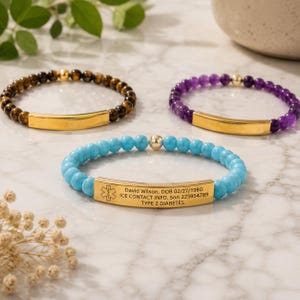 May include: Three beaded bracelets featuring gold-tone metal bars. One bracelet has turquoise beads, one has purple beads, and one has brown tiger eye beads. The turquoise bracelet is engraved with a medical symbol and personal information.